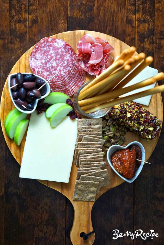 Fall-themed charcuterie board that includes a Goat Cheese Log, Gruyere Cheese, Ossau-Iraty Cheese, Salami, Prosciutto, Sourdough crackers, Breadsticks, Kalamata Olives, Sundried Tomatoes, Apples, Pistachios, and Cranberries.