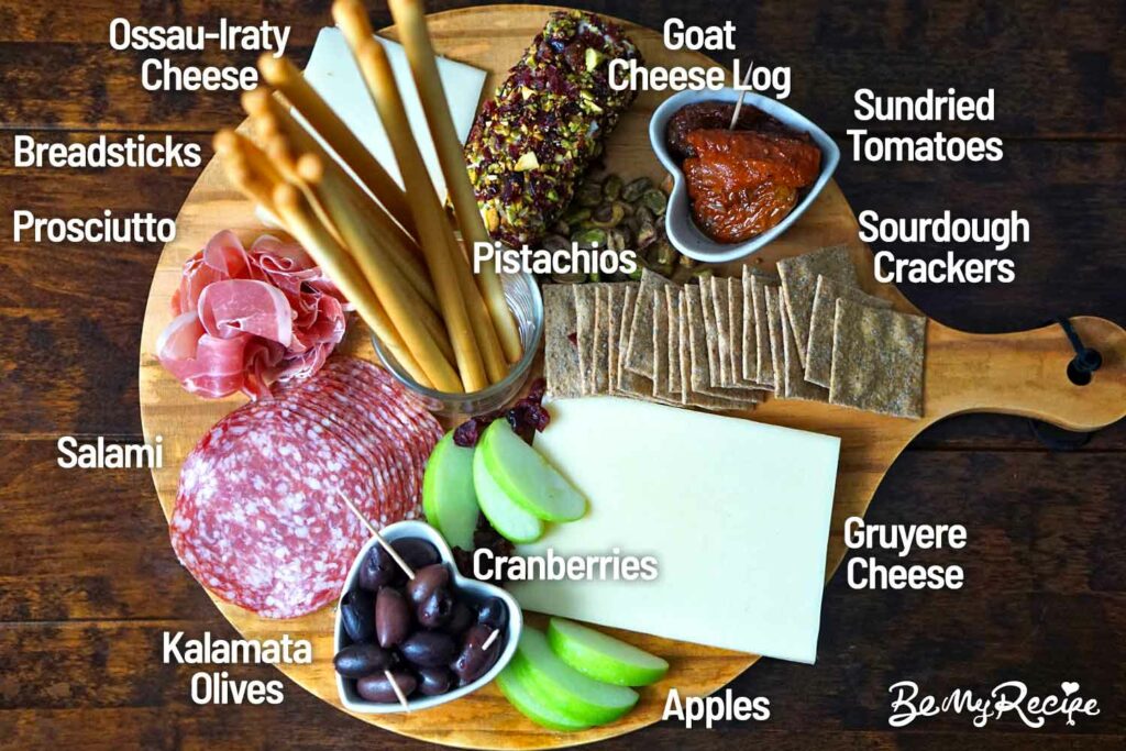 Charcuterie board that includes a Goat Cheese Log, Gruyere Cheese, Ossau-Iraty Cheese, Salami, Prosciutto, Sourdough crackers, Breadsticks, Kalamata Olives, Sundried Tomatoes, Apples, Pistachios, and Cranberries.