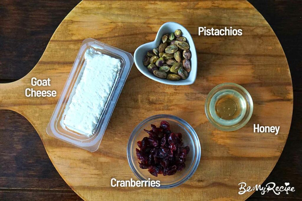 Ingredients on a board (featuring a goat cheese log, roasted pistachios, honey, and dried cranberries.