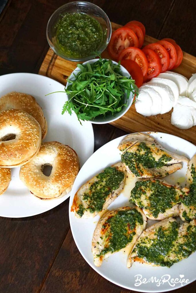 Assembling the bagel (pan-fried chicken with homemade pesto, sliced mozzarella, arugula (or rocket), sliced tomatoes, and toasted sesame bagels).