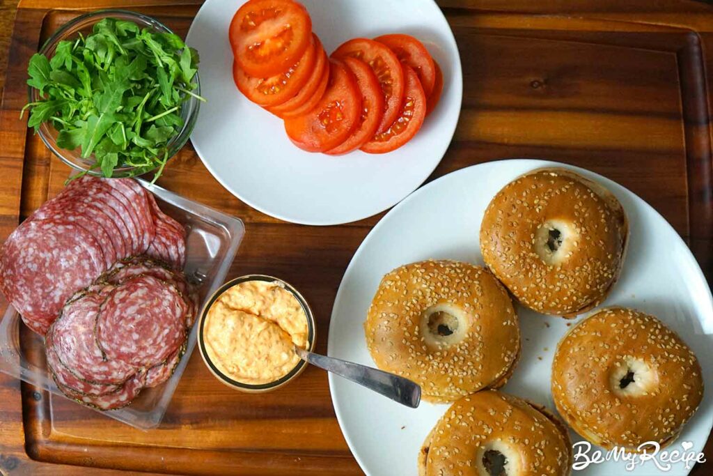Bagel assembly board (toasted bagels, salami, roasted pepper cream cheese, arugula, and tomatoes).