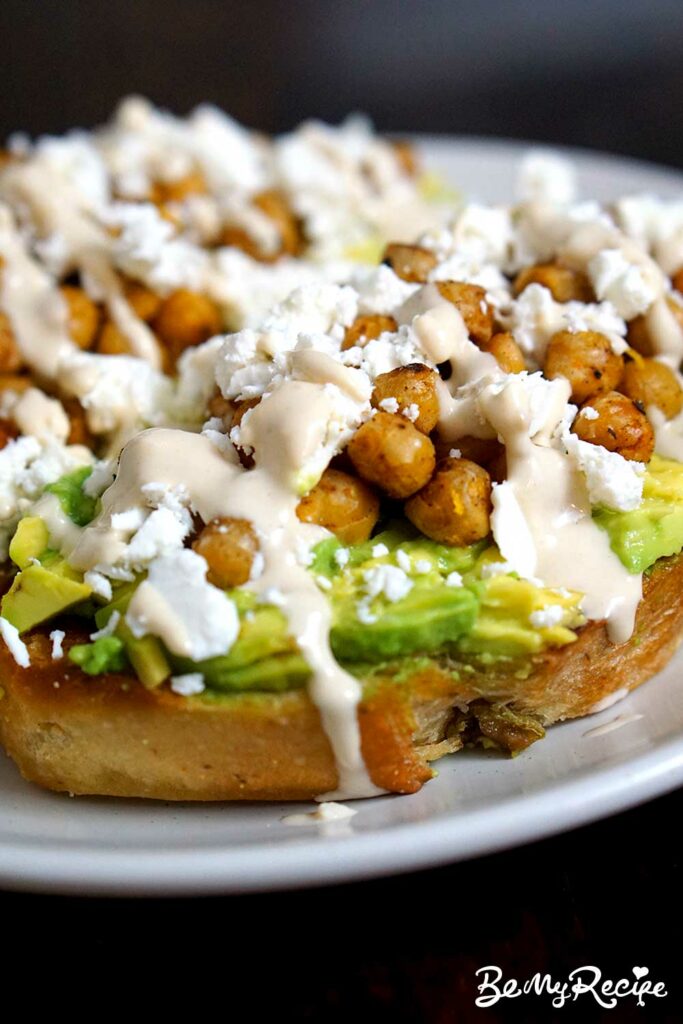 Toasted bread slices with mashed avocado, warm chickpeas, feta, and tahini sauce