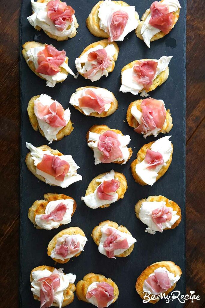 Topping the toasted baguette slices with mozzarella and prosciutto