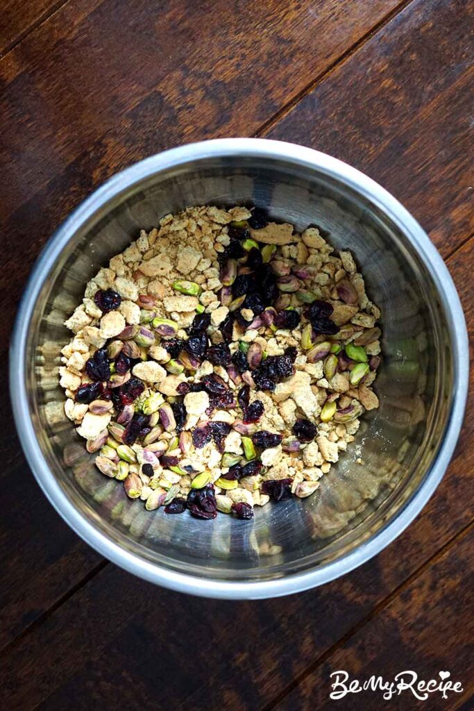 top down view of crushed biscuit, cranberries, and nuts in a metal mixing bowl against a dark wooden surface