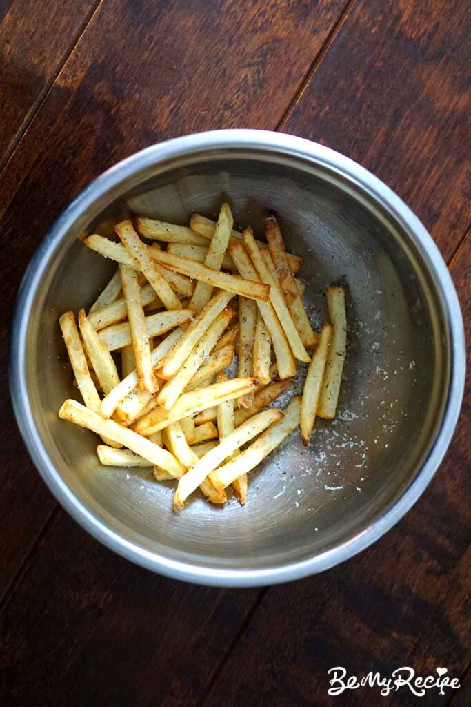 potato fries being tossed in a metal mixing bowl with thyme, parmesan, salt, and pepper