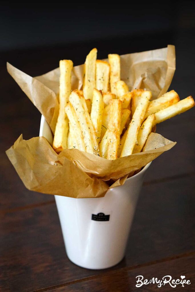 air fryer fries with parmesan, garlic, and thyme in a round container displayed upright wrapped in paper on a wooden table