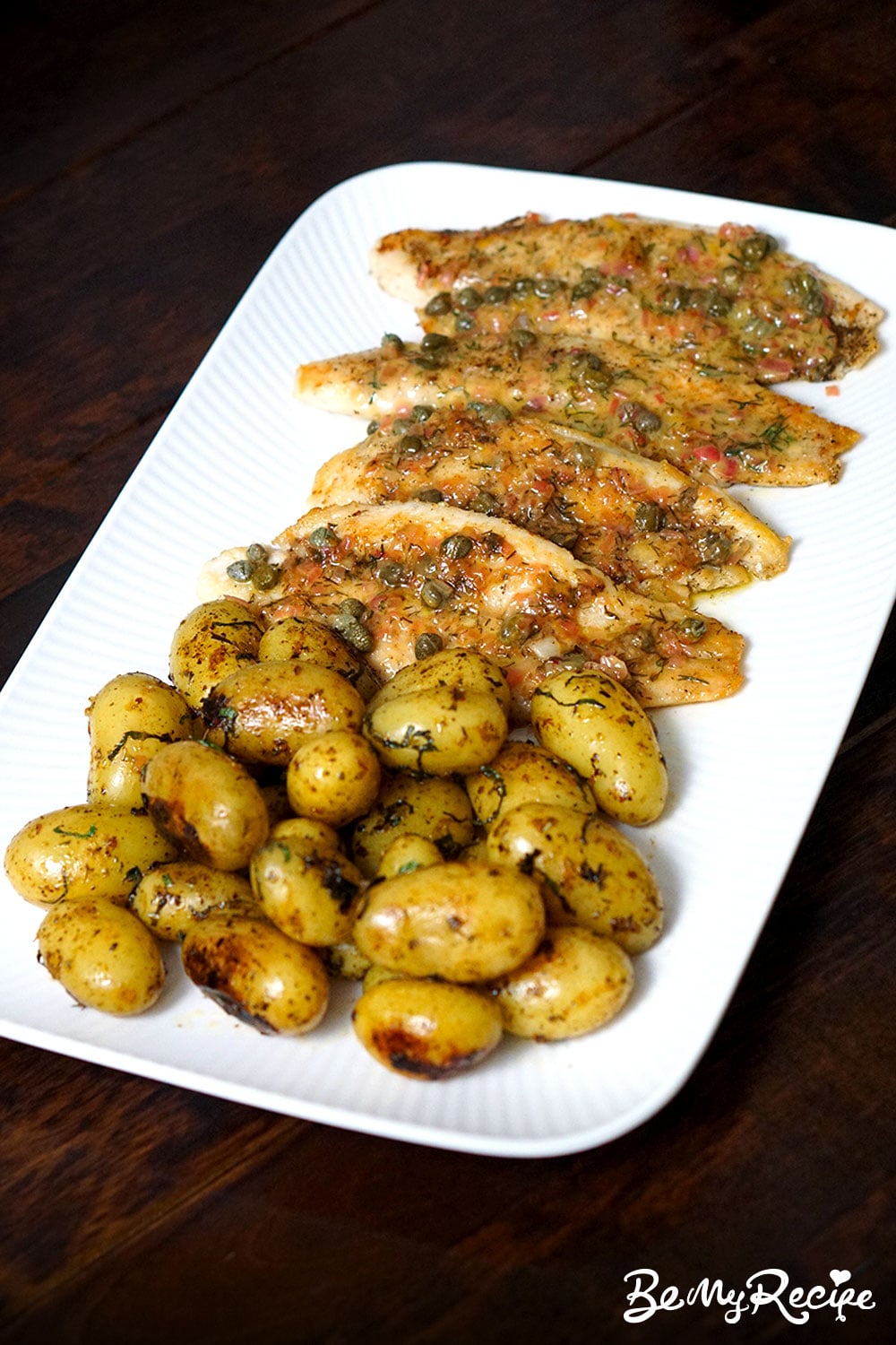 overhead angled view of fish fillets with lemon butter sauce and new potatoes laid out on a white rectangular platter against a dark background