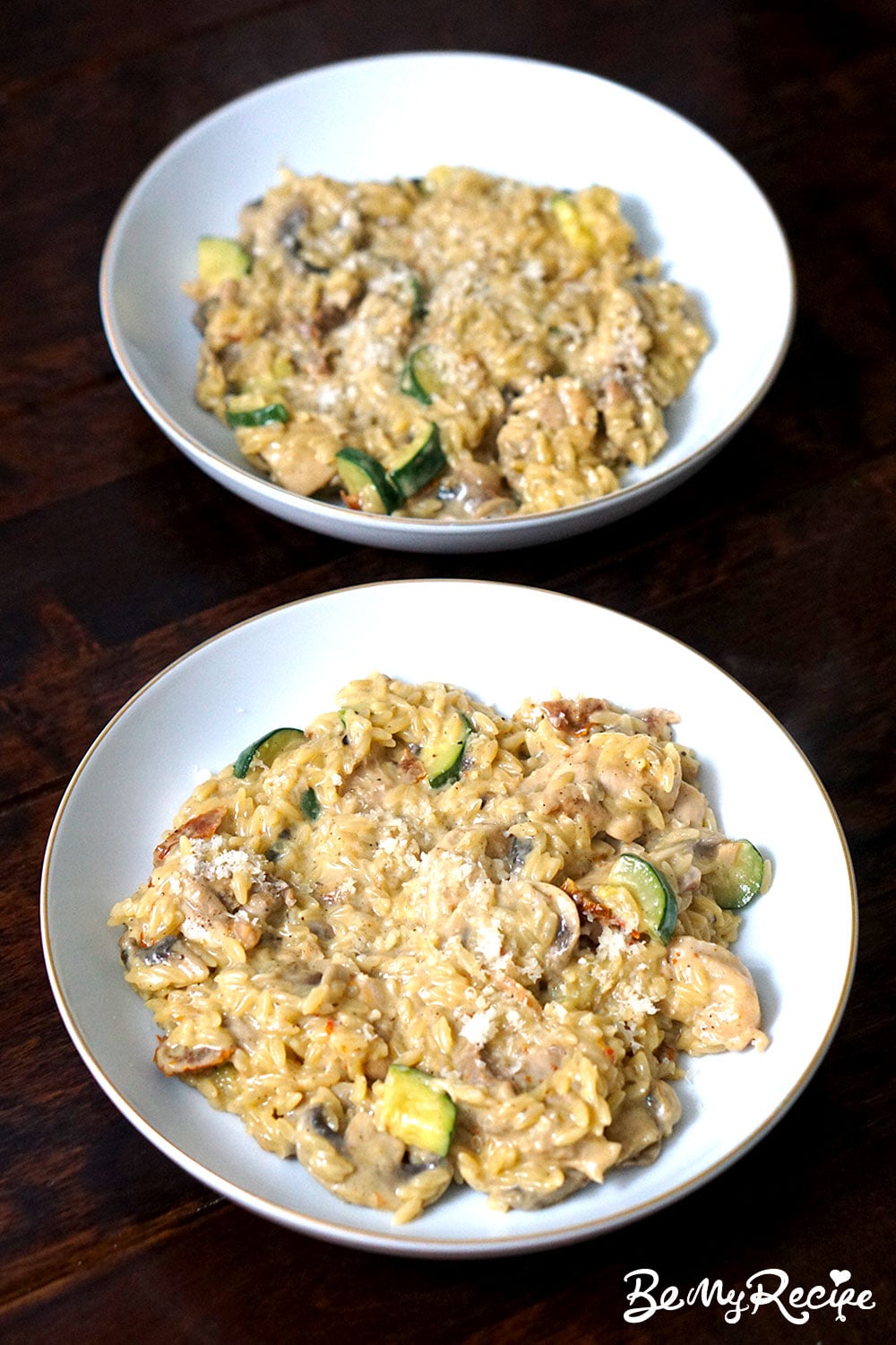 overhead angled view of two white bowls of chicken orzo, mushrooms, zucchini, and sundried tomatoes, topped with grated parmesan, displayed on a dark wooden background