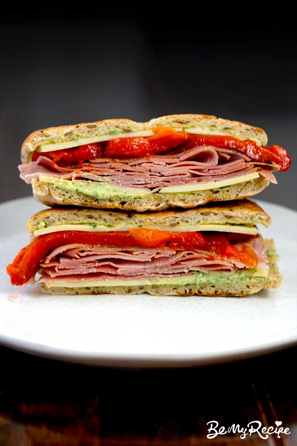 cross section of ham and cheese sandwich with pesto yogurt and roasted peppers on a white plate against a black background