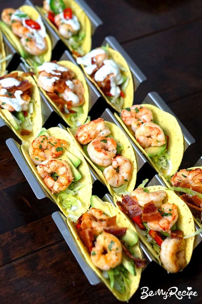 angled view of 12 shrimp tacos in rows of 3, with the central tacos having no toppings, the row at the front topped with crispy bacon, and the two rows behind topped with bacon and/or creme fraiche