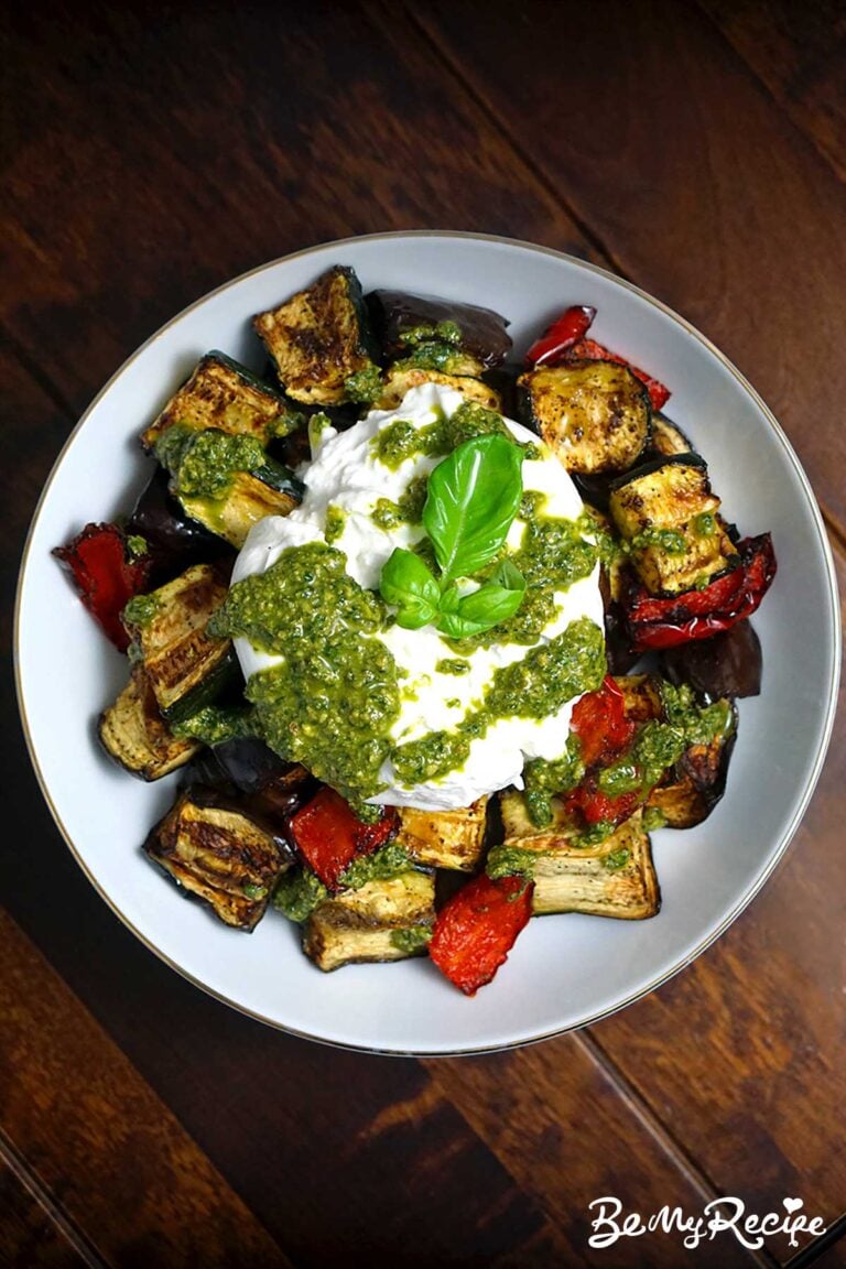 top down view of air-fryer veggies topped with burrata and fresh basil pesto in a white bowl against a dark wooden surface