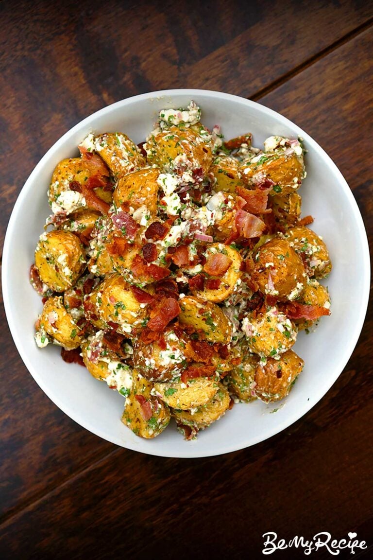 overhead view of warm potato salad with crispy bacon and crumbled feta served in a white bowl against a dark wooden background